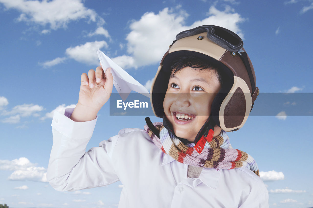 Close-up of happy boy holding paper airplane while wearing helmet against cloudy sky