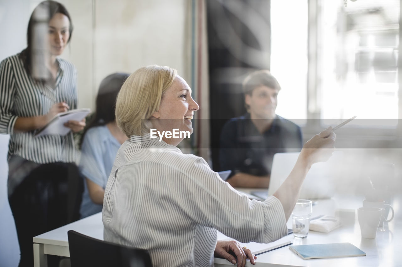 Smiling businesswoman pointing while sitting with colleagues in board room during meeting