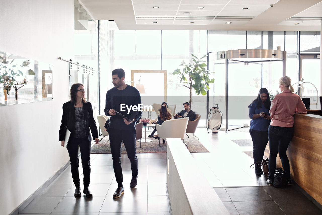 Mature businesswoman discussing with male colleague while walking in lobby with coworkers working in background