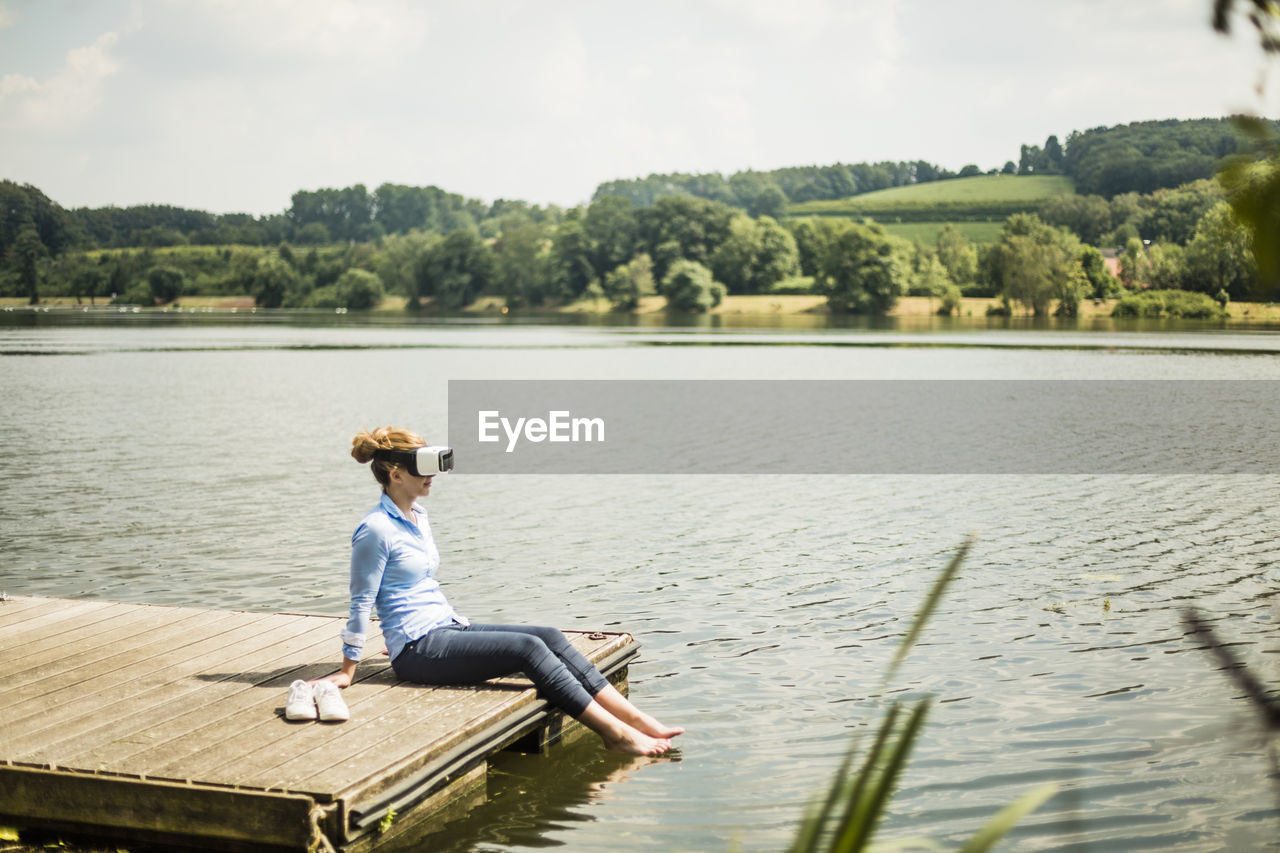 Woman with vr glasses sitting on jetty at a lake with feet in water