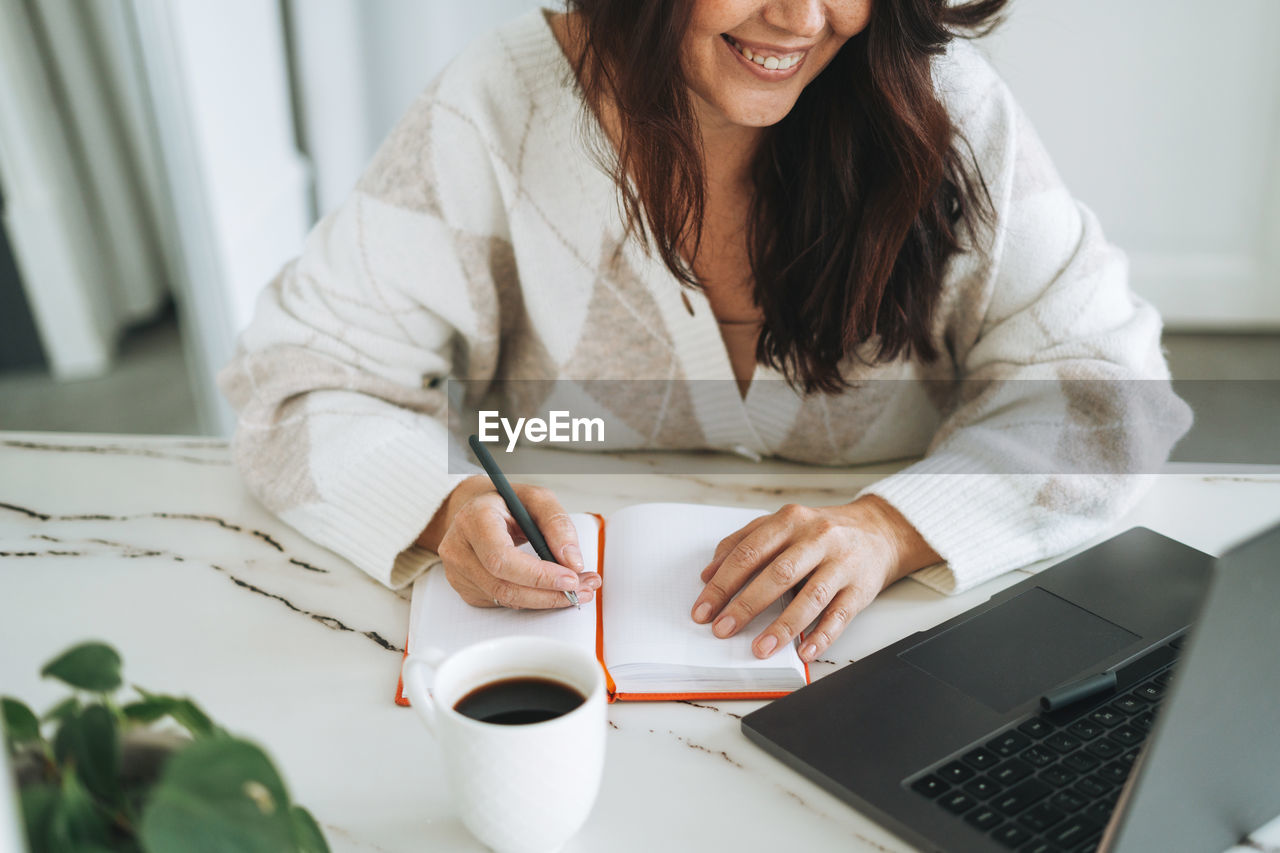 Smiling brunette woman with long hair in white cardigan working on laptop in office