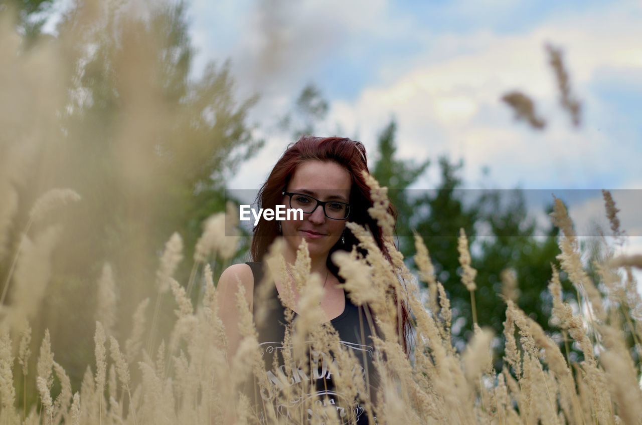 PORTRAIT OF SMILING YOUNG WOMAN STANDING BY TREE ON FIELD AGAINST SKY