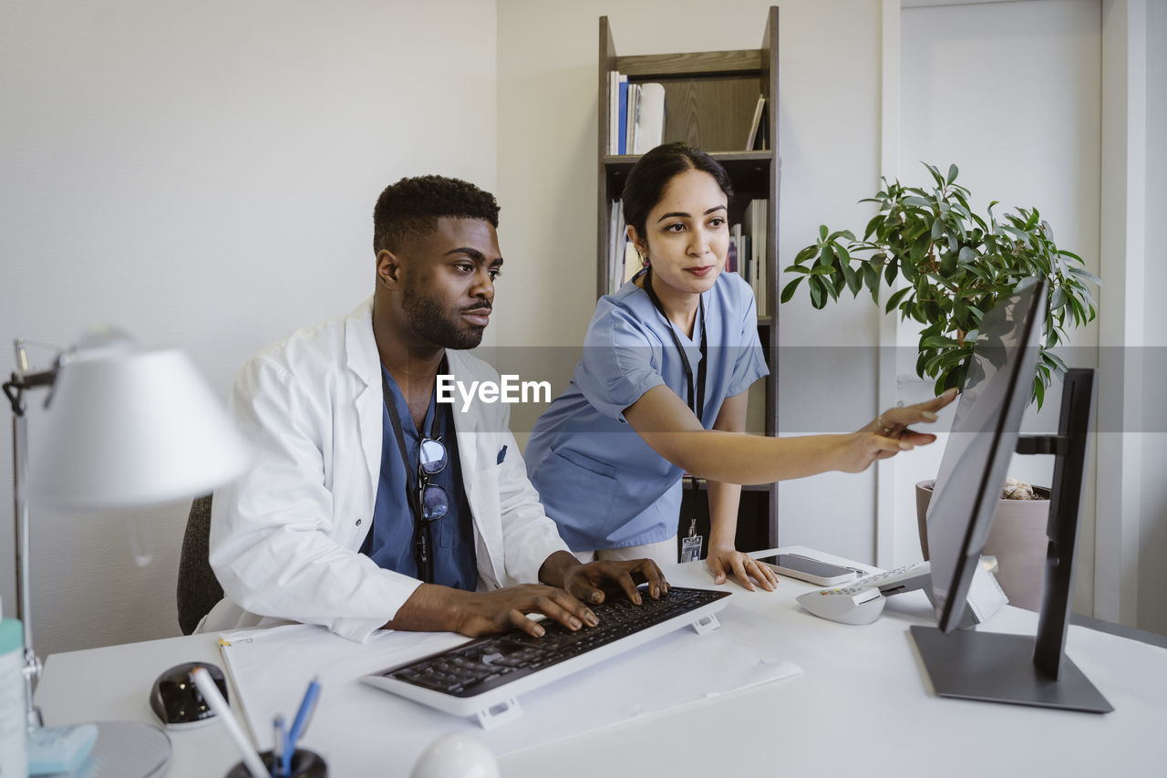 Doctor discussing with male coworker working on computer at desk in clinic