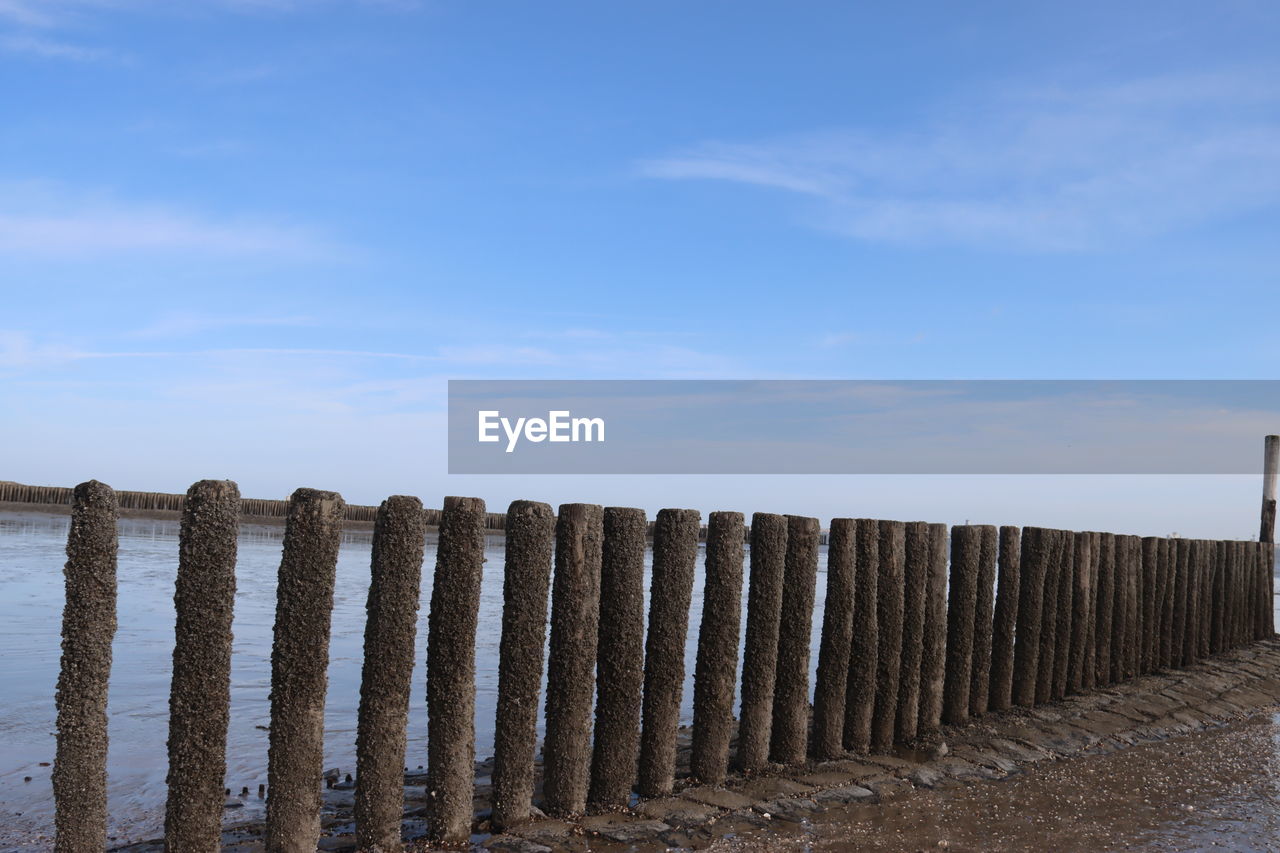 sky, cloud, fence, nature, no people, water, land, protection, wall, security, wood, day, beach, sea, tranquility, post, scenics - nature, in a row, outdoors, tranquil scene, wooden post, architecture, home fencing, sand, coast, environment, beauty in nature, blue, horizon, built structure