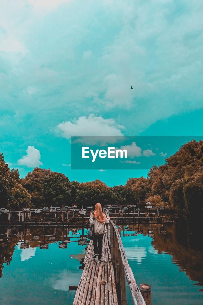 Rear view of woman walking on footbridge over river against sky