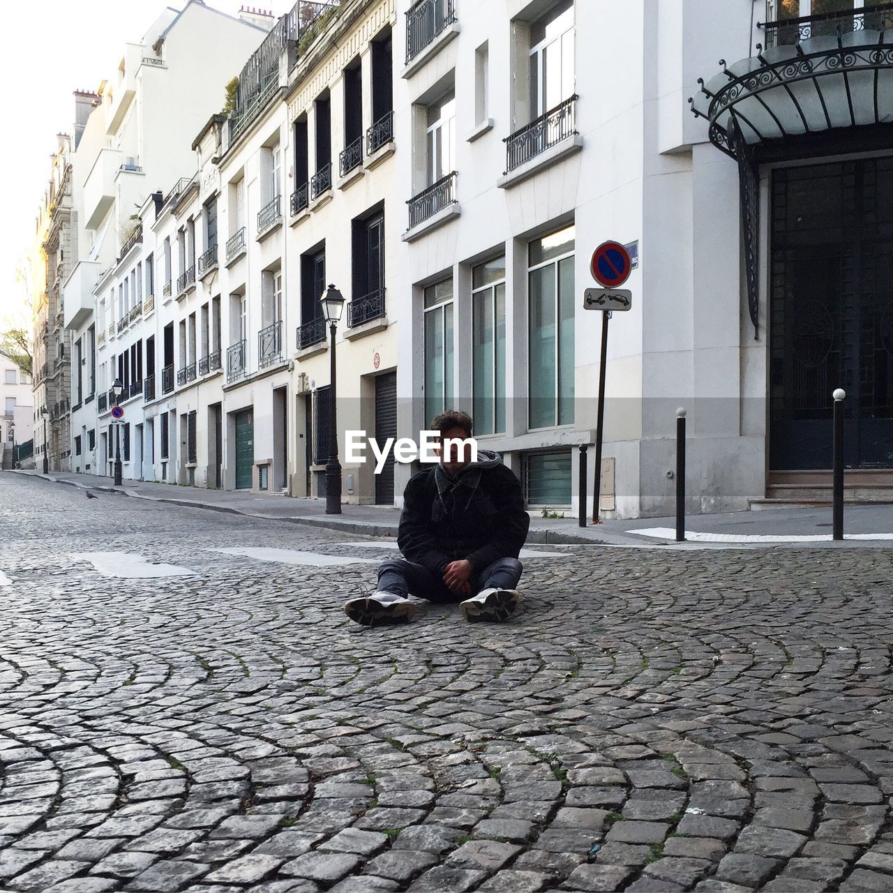 Full length portrait of man sitting on cobbled street by buildings