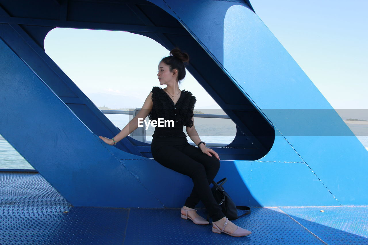 Full length of young woman sitting on metallic built structure on ship in sea against blue sky