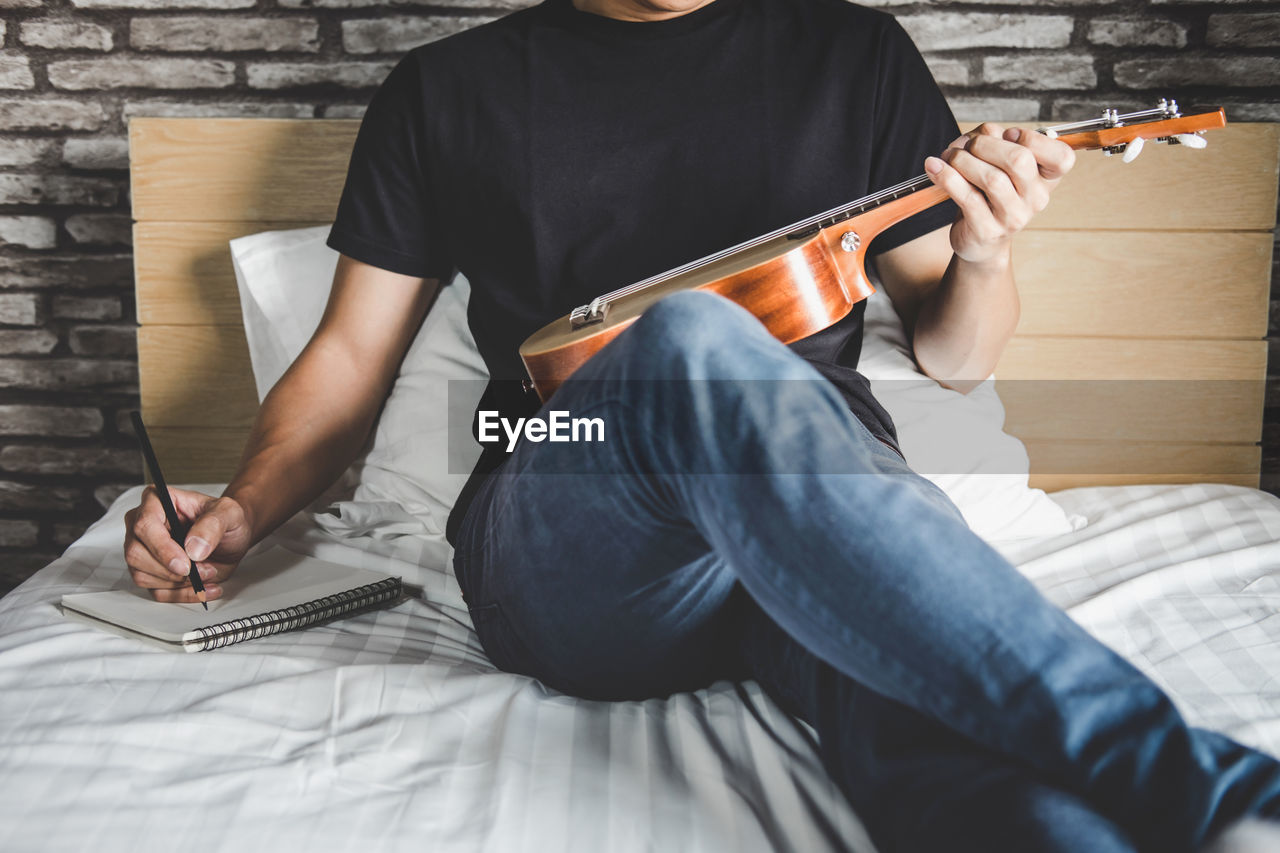 Midsection of man holding musical instrument while sitting on bed at home