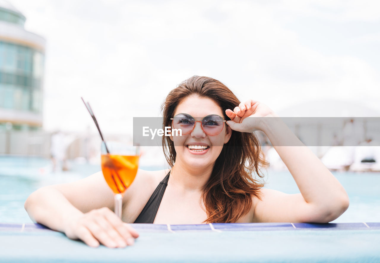 Stylish woman plus size body positive in black swimsuit with glass of cocktail in pool