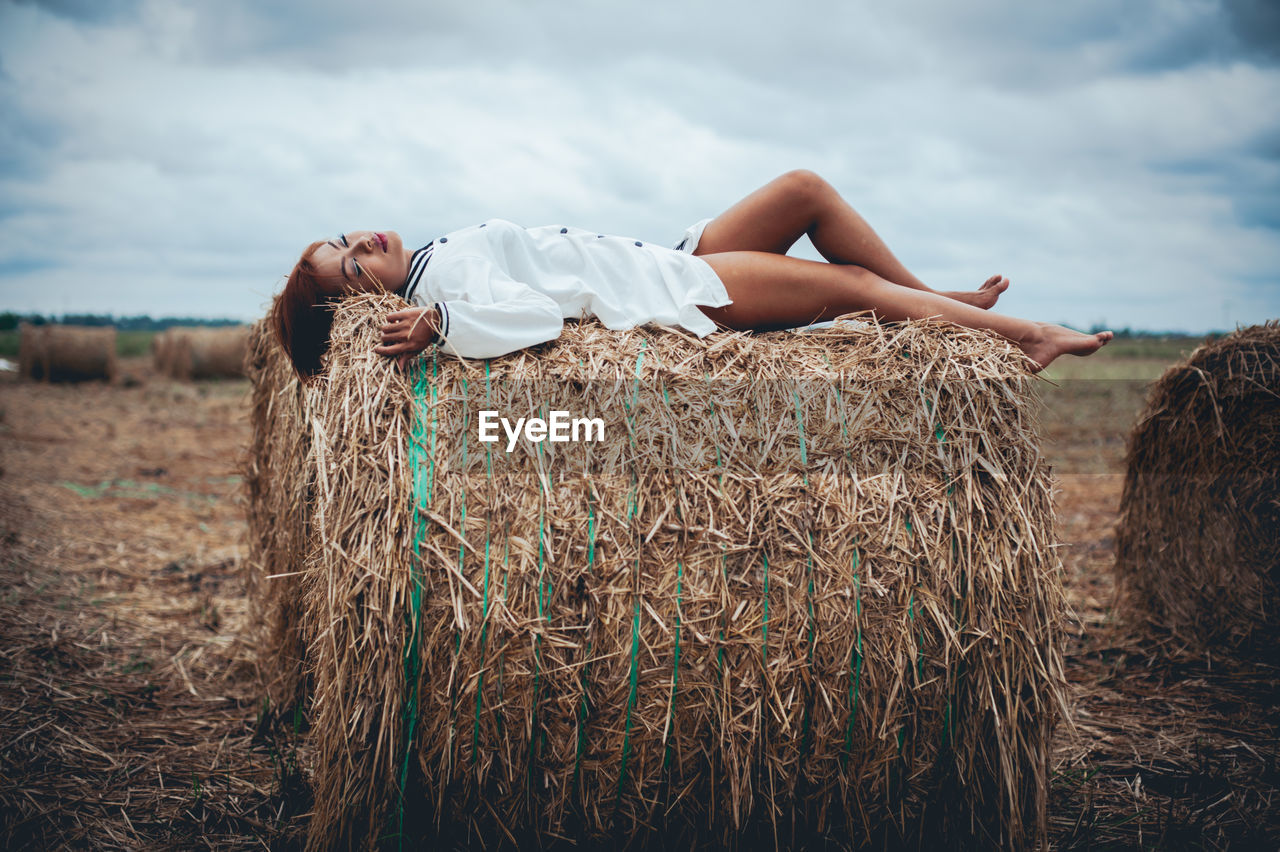 Side view of young woman sleeping on hay bale against cloudy sky at farm