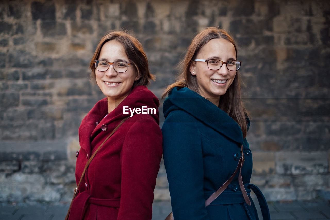 Portrait of smiling sisters standing back to back against stone wall