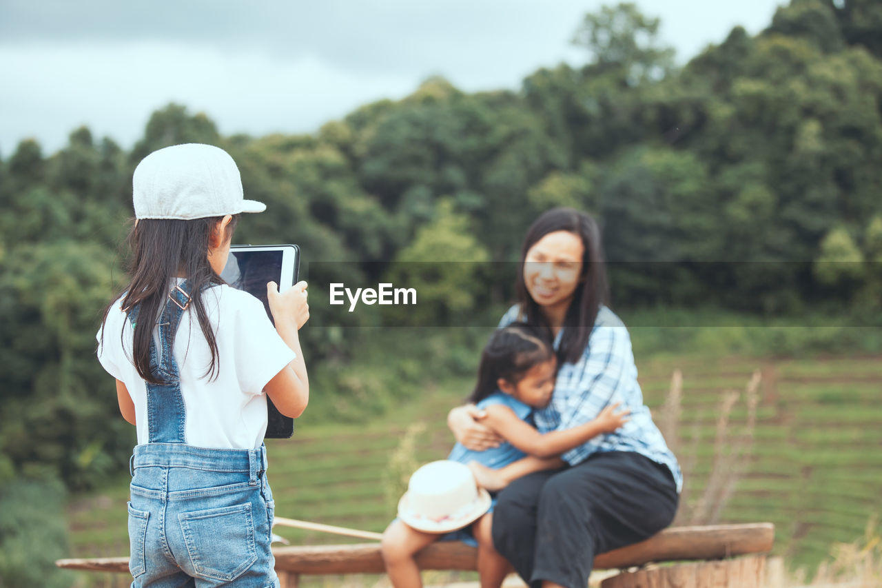 Rear view of girl photographing mother and daughter in park