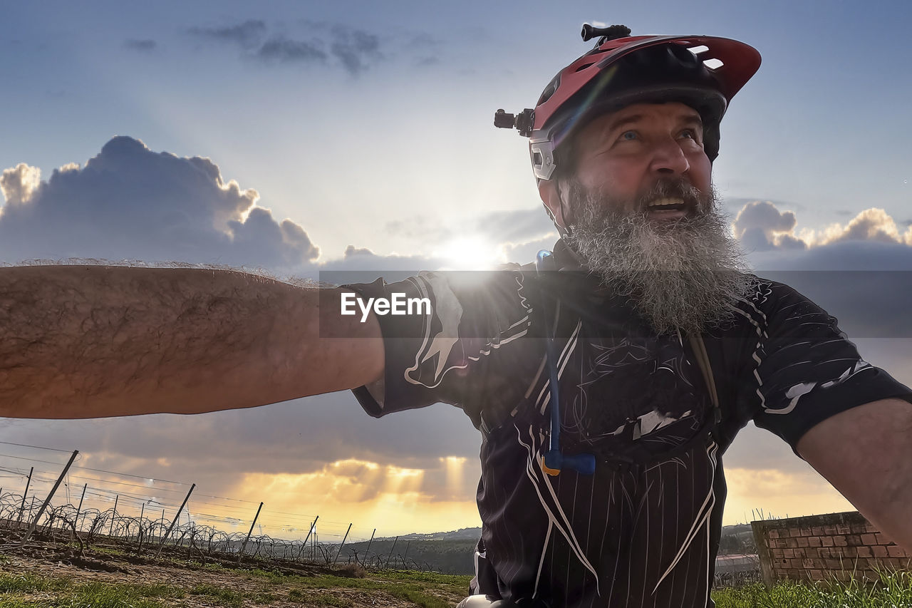 adult, sky, one person, nature, men, facial hair, beard, cloud, sunset, mountain, sunlight, sports, leisure activity, lens flare, landscape, lifestyles, clothing, portrait, activity, sun, person, back lit, outdoors, human face, rural scene, land, beauty in nature, adventure, waist up, smiling, standing, emotion, hat, mature adult, technology, grass, happiness