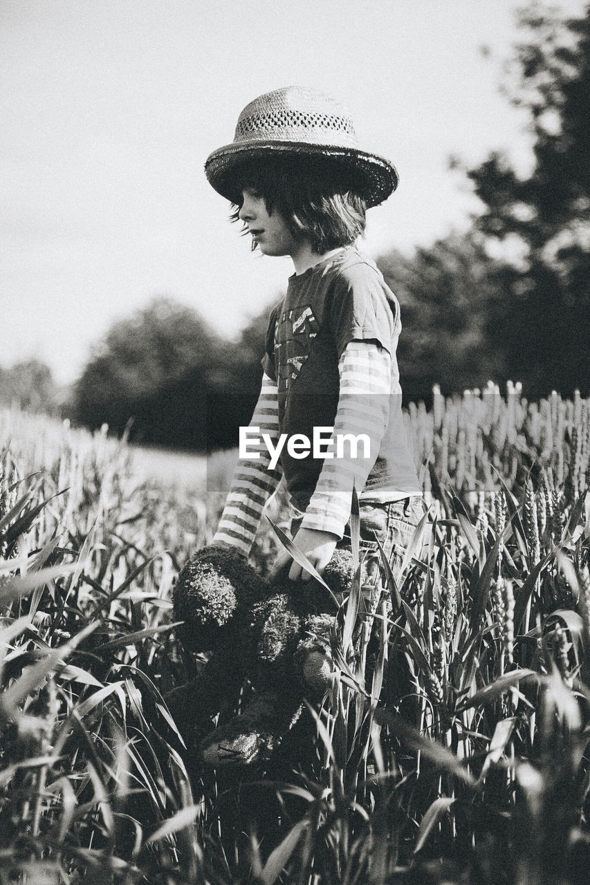 Boy holding teddy bear walking amidst wheat field against sky