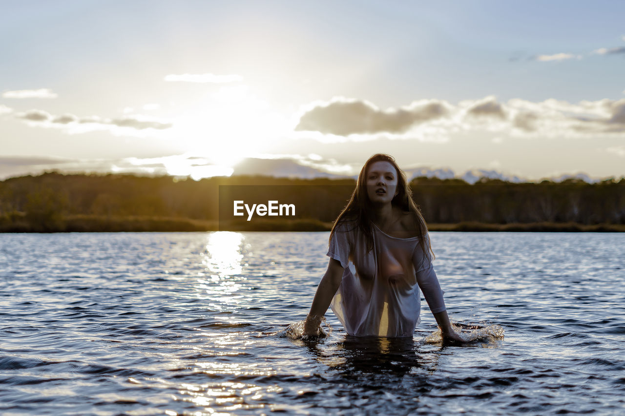 Portrait of young woman standing in lake against sky during sunset