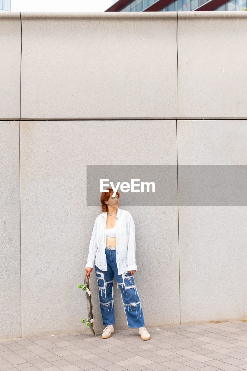 Full body of female in casual wear with skateboard looking away while standing on gray background in city