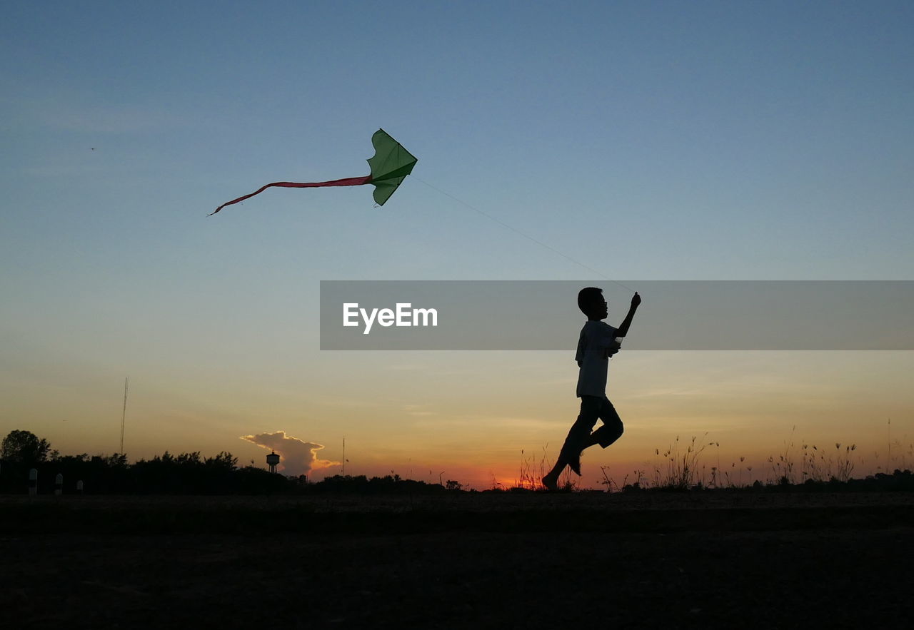 Boy flying kite while running on field during sunset