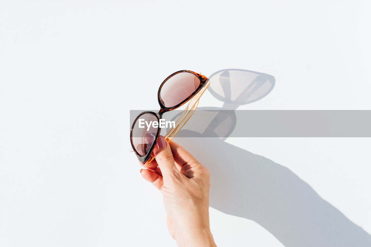 From above top view of cropped anonymous female hands holding trendy sunglasses with brown lens and frame over white background with shadow