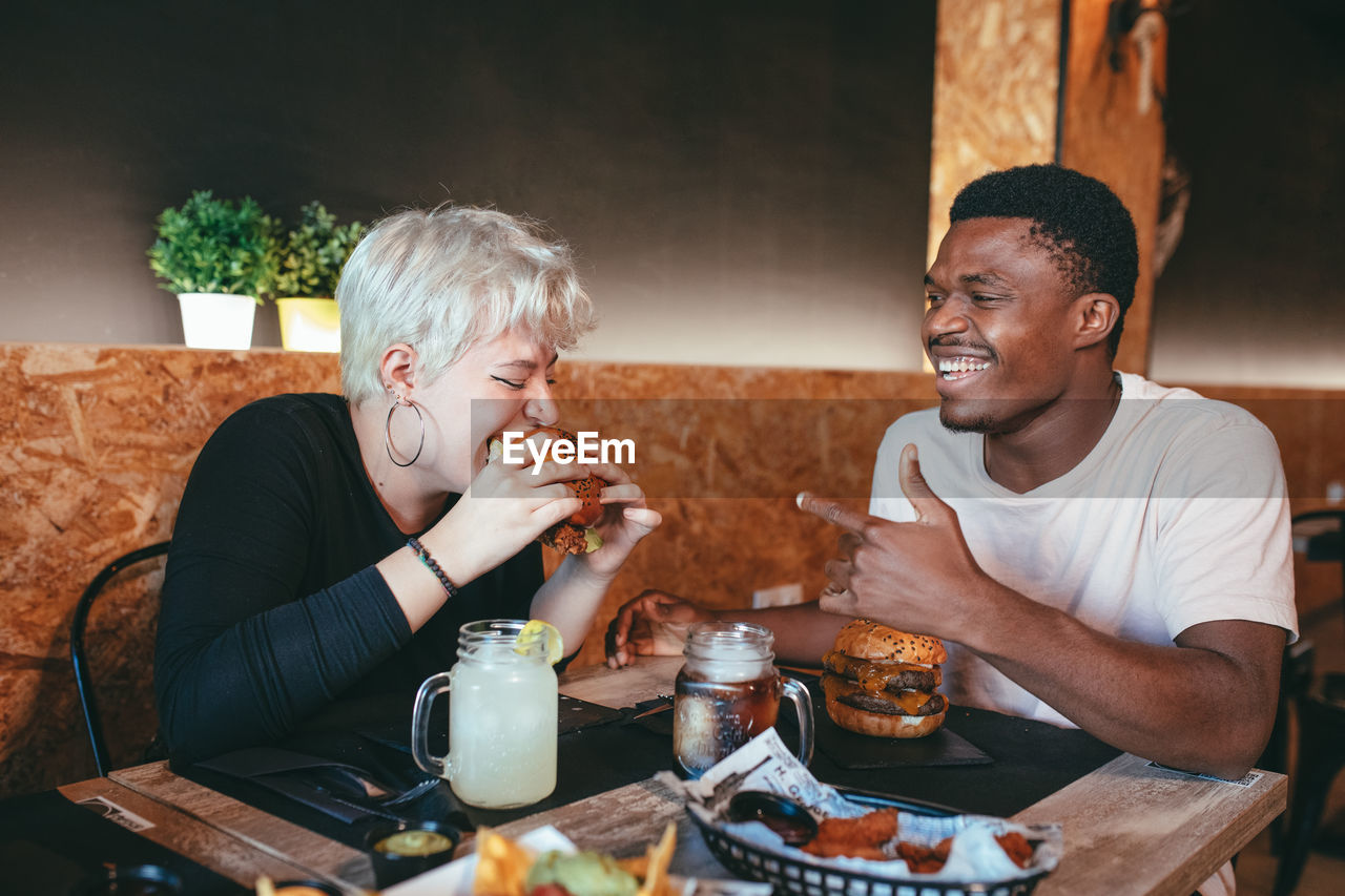 Cheerful woman and black male friend sitting in fast food cafe having fun while eating burgers