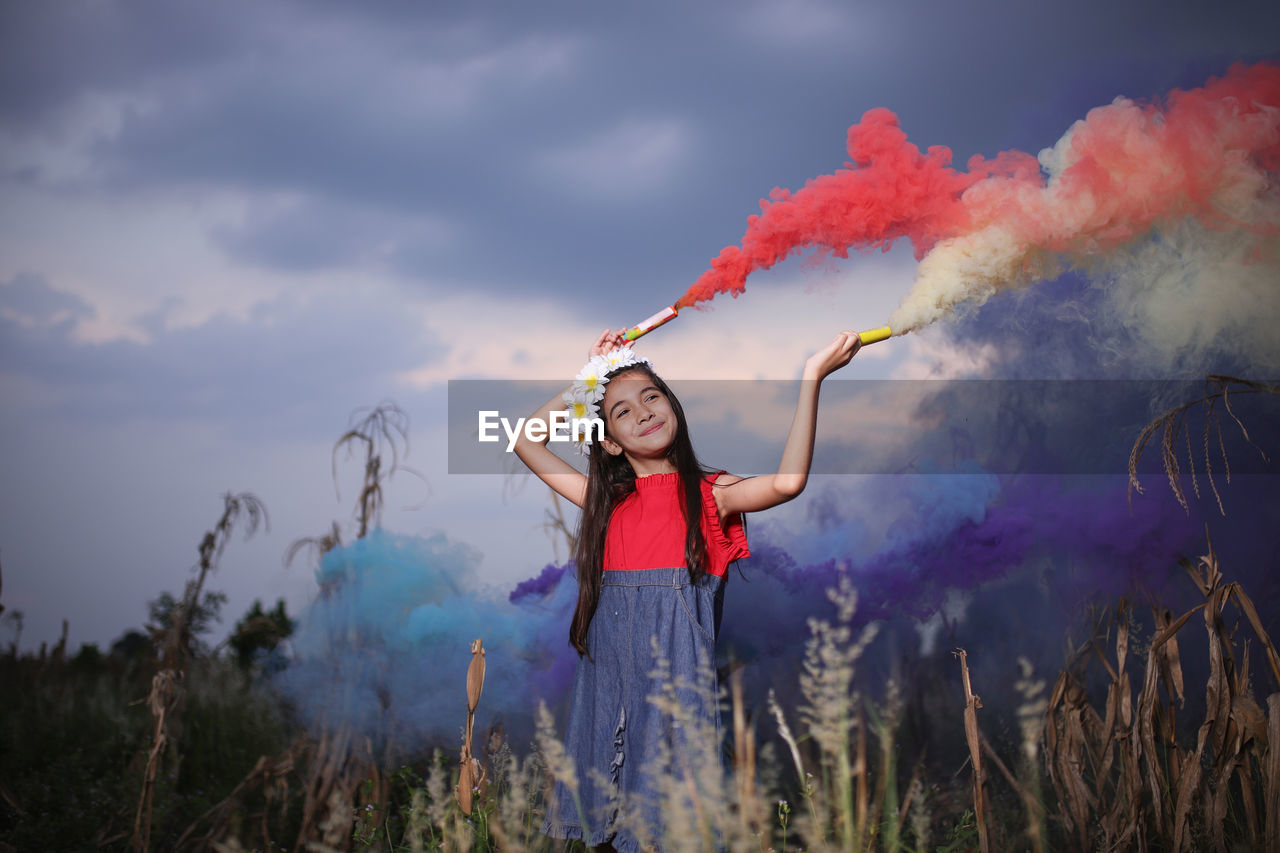 Smiling cute girl holding distress flares while standing against sky at dusk