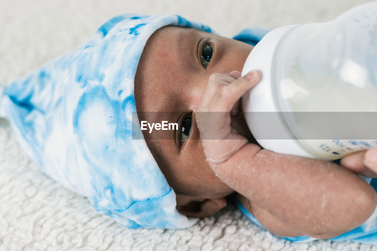 Cropped unrecognizable mother feeding tranquil adorable newborn baby with milk bottle wrapped on blue cozy blanket