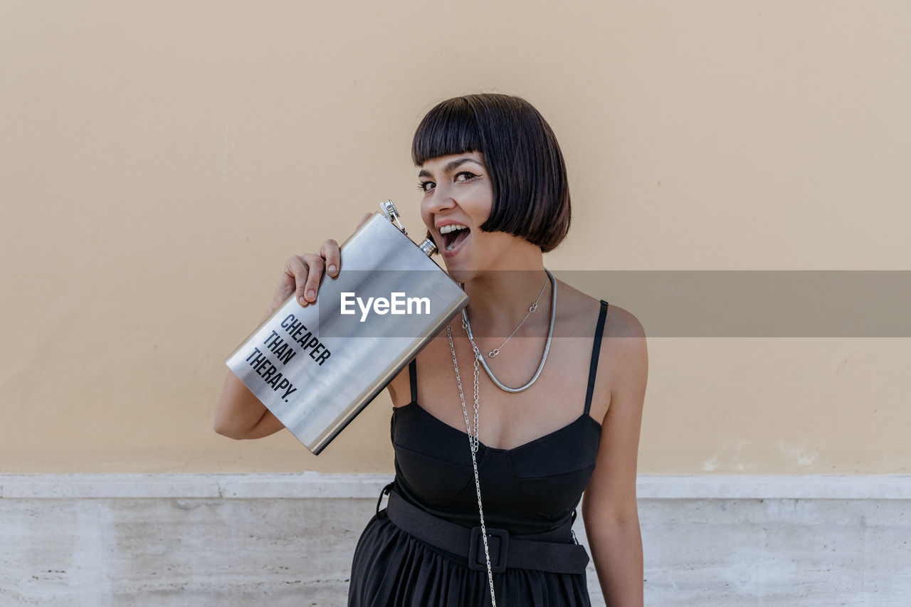 Portrait of happy young woman wearing elegant black jumpsuit, holding large liquor flask