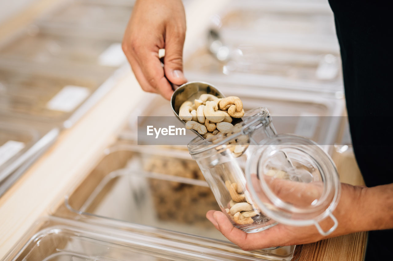 Side view of anonymous female pouring cashews inside glass jar in local store