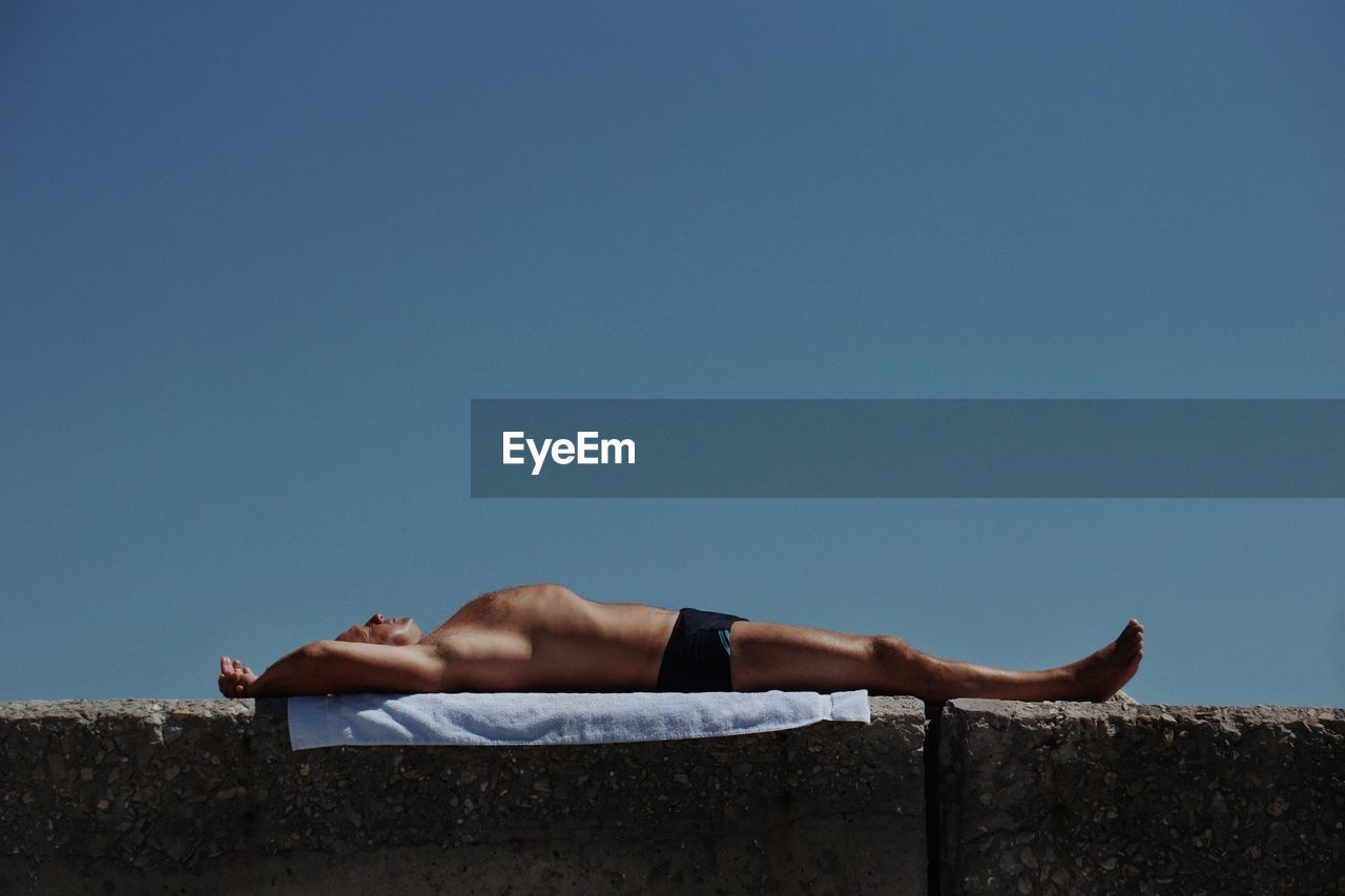 Shirtless man relaxing on retaining wall against clear blue sky