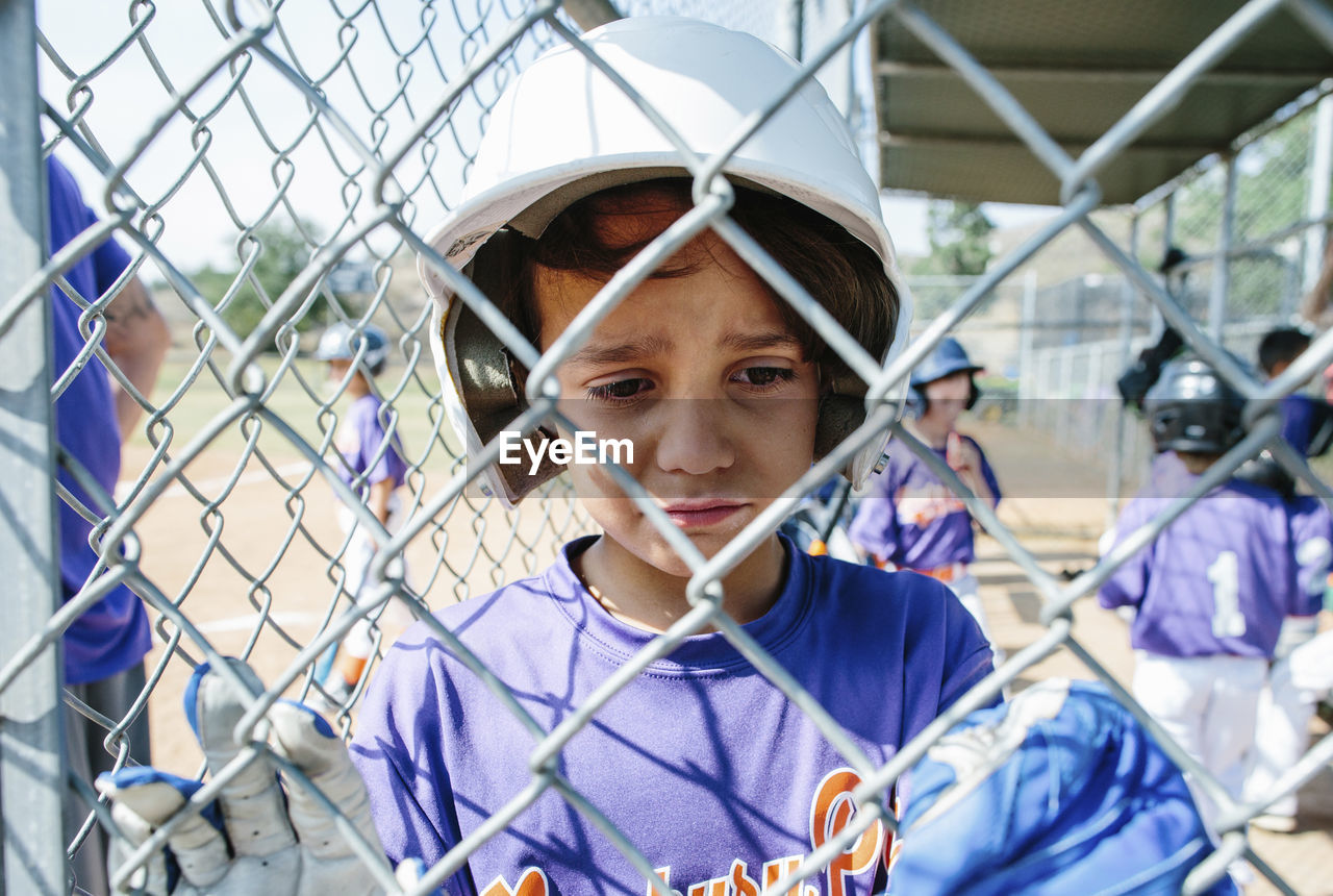 Close-up of sad boy standing in sports dugout seen through chainlink fence