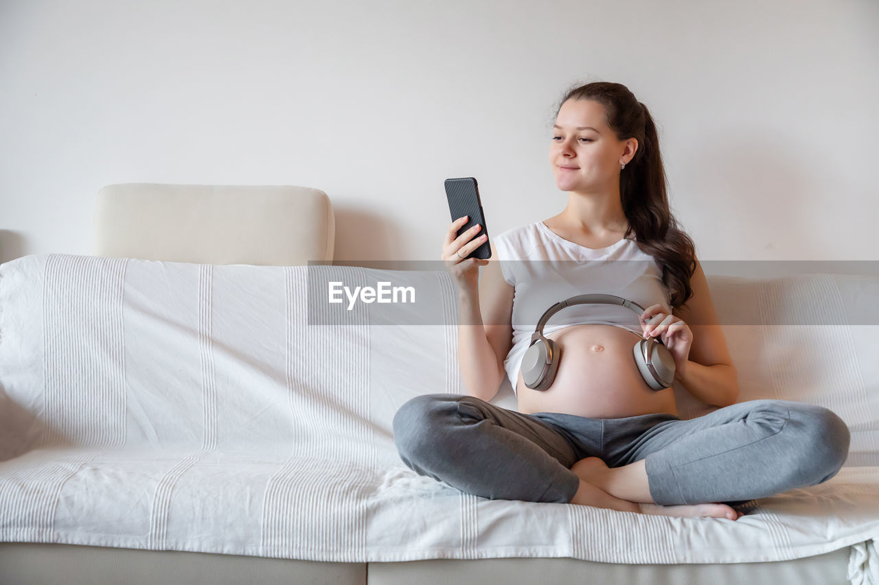 Young pregnant woman holding headphones on stomach while sitting on sofa at home