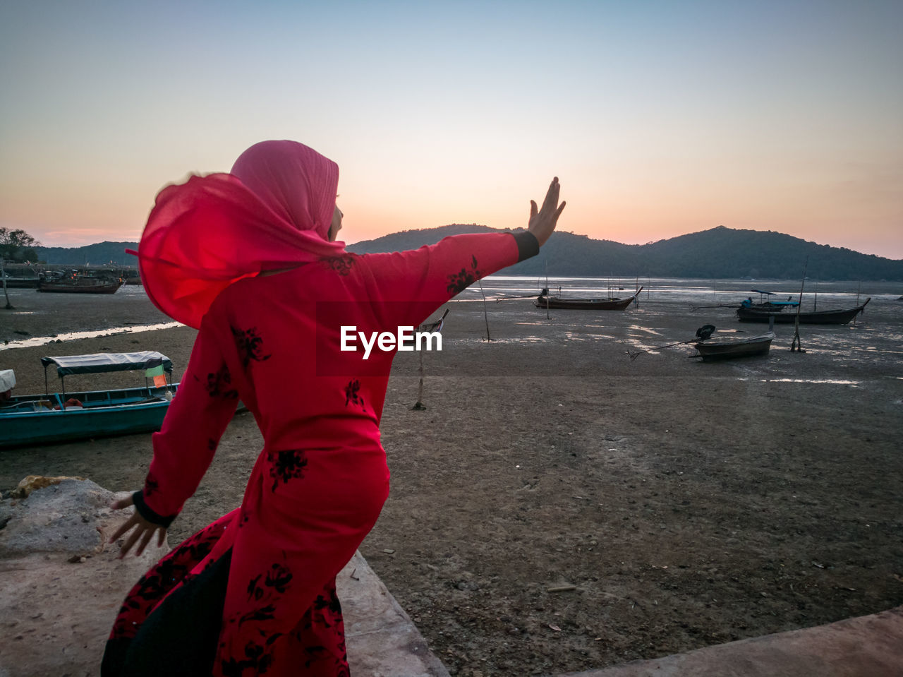 Rear view of girl gesturing while standing at beach against sky during sunset