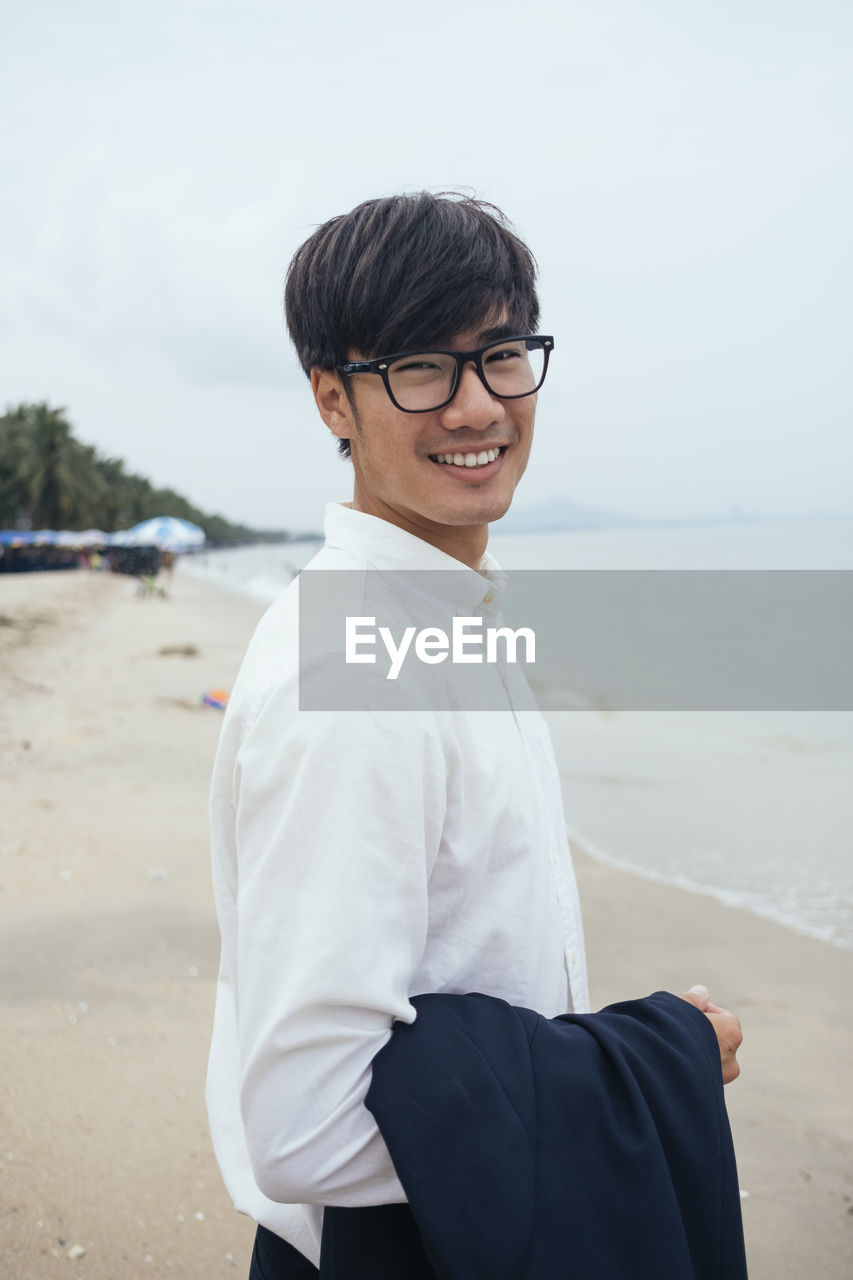 PORTRAIT OF SMILING YOUNG MAN STANDING AT BEACH