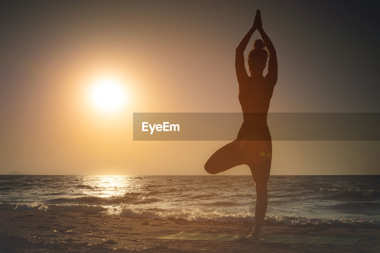 Full length rear view of a silhouette of a woman standing on one leg while practicing tree pose in