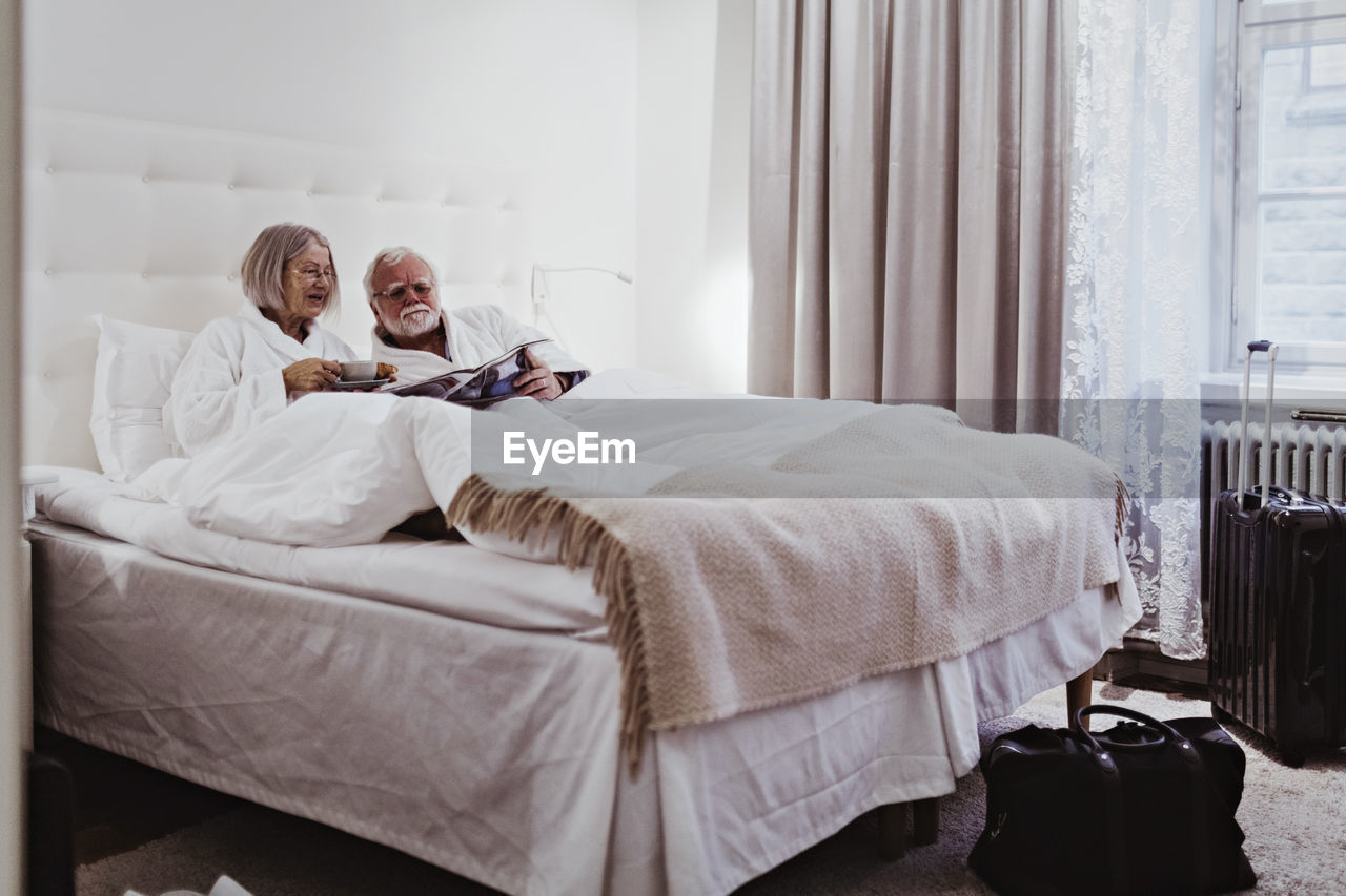 Senior woman reading newspaper while having coffee by man relaxing on bed in hotel