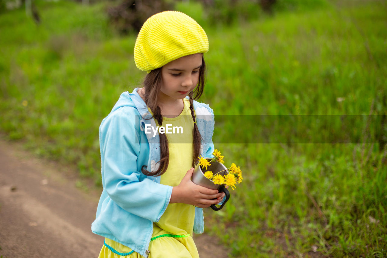 A little girl walks along a path near a green lawn with a bouquet of yellow dandelions in an mug