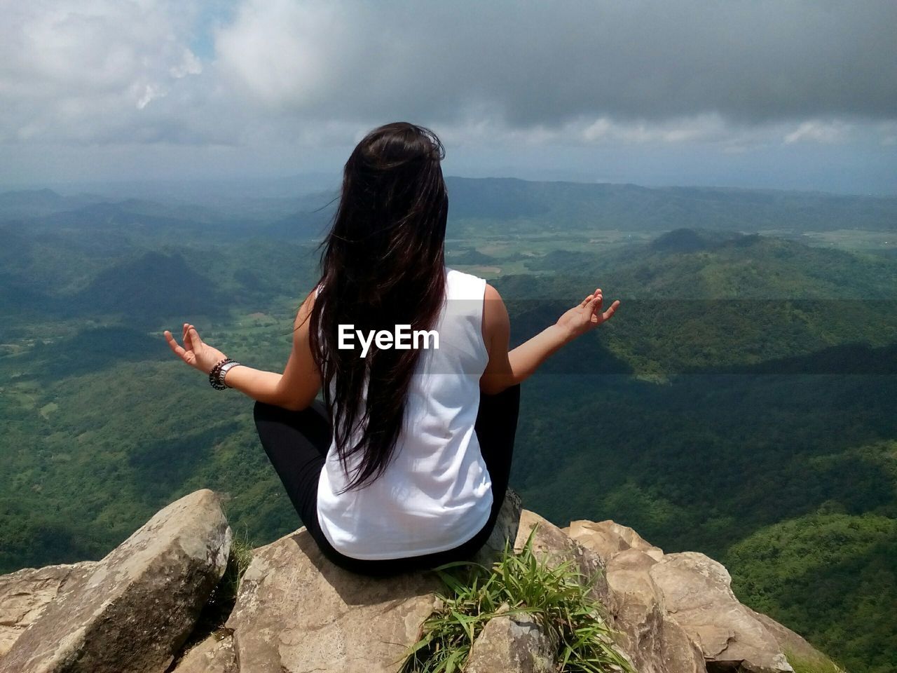 Rear view of woman performing yoga while sitting on rocks against cloudy sky