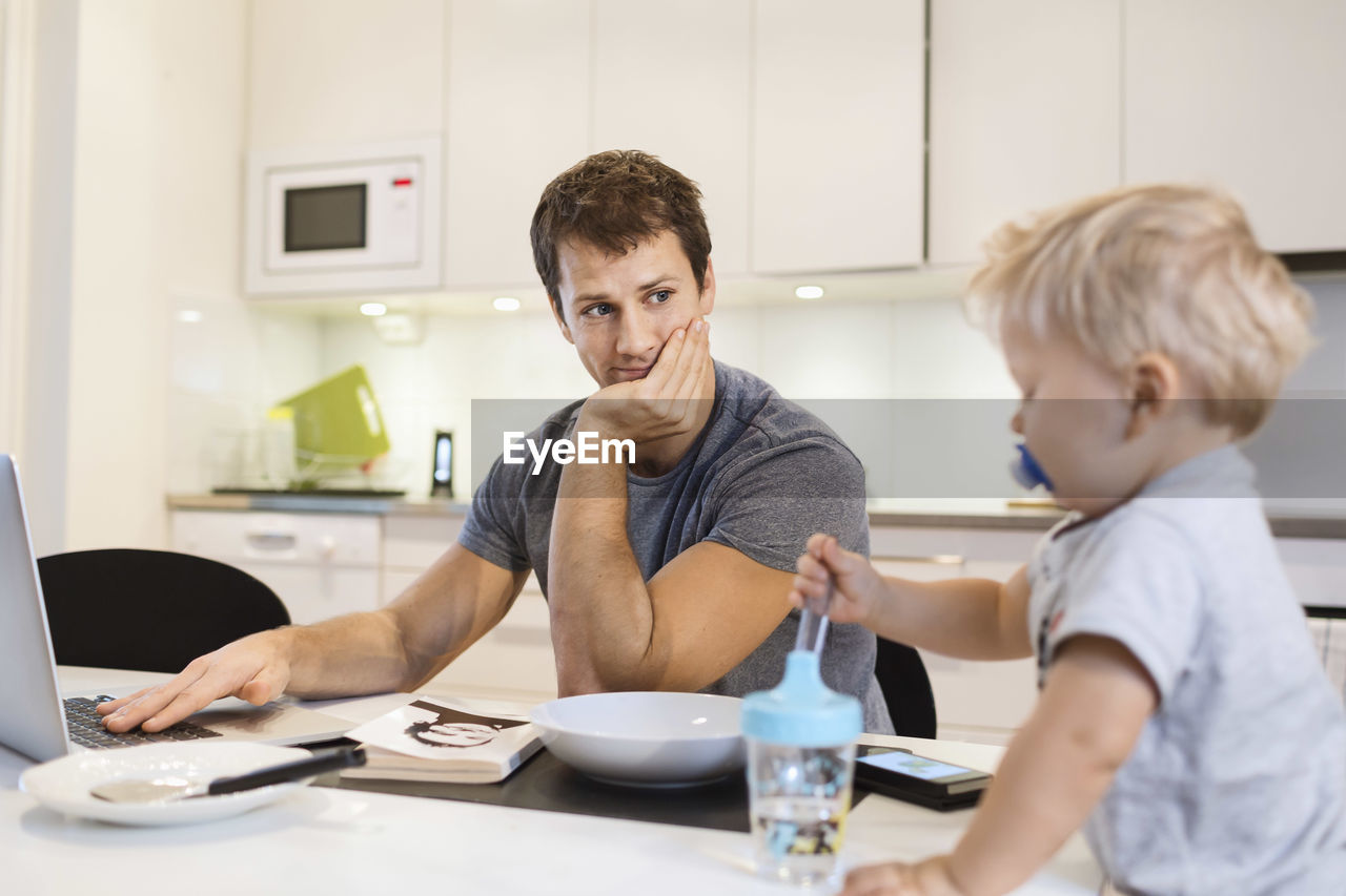 Father looking at baby boy while using laptop in kitchen