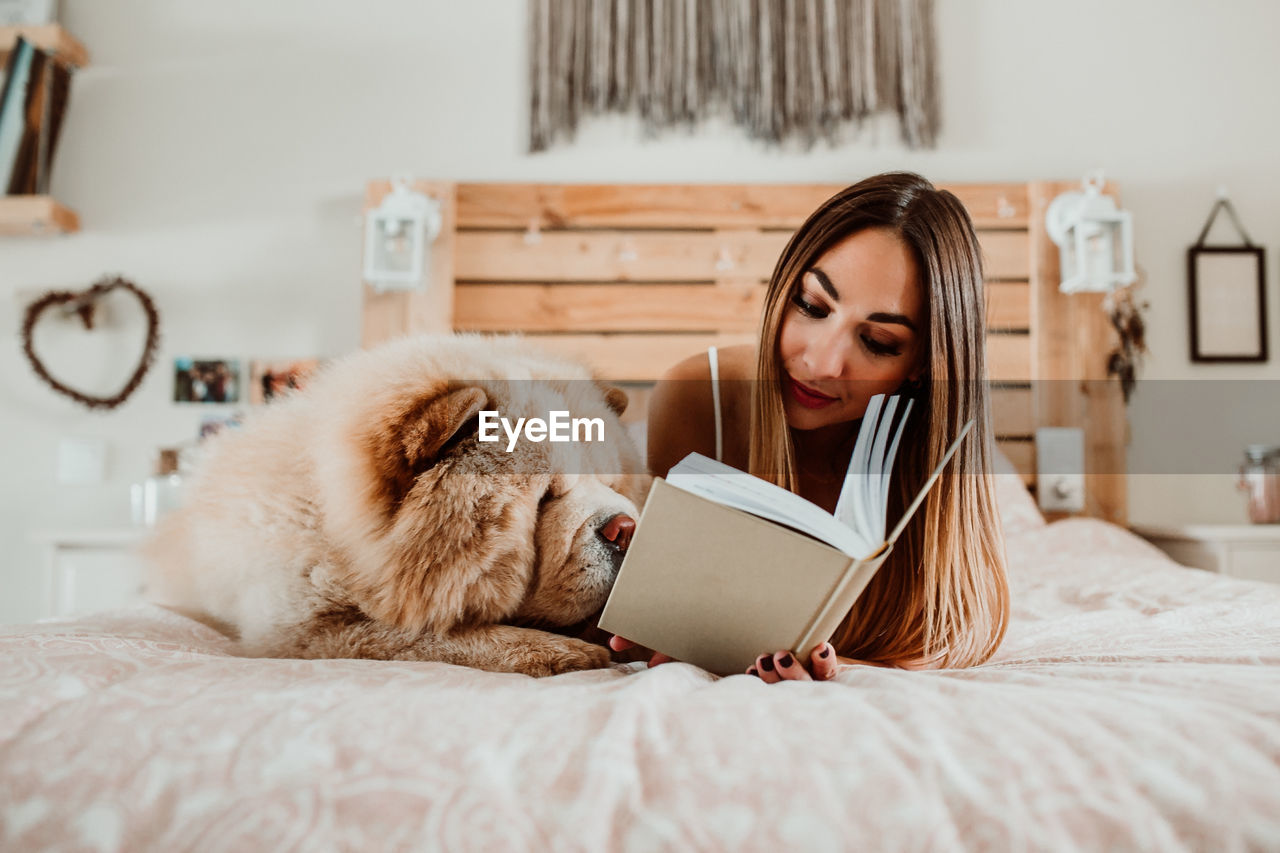 Young woman reading book while lying with dog on bed