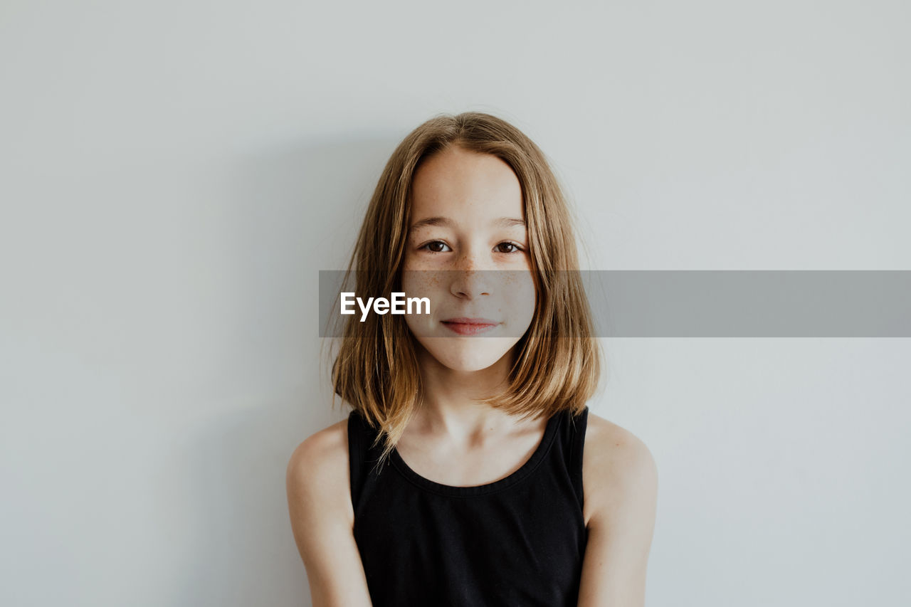 Calm adorable teenage girl with freckles in casual top standing against white background and looking at camera