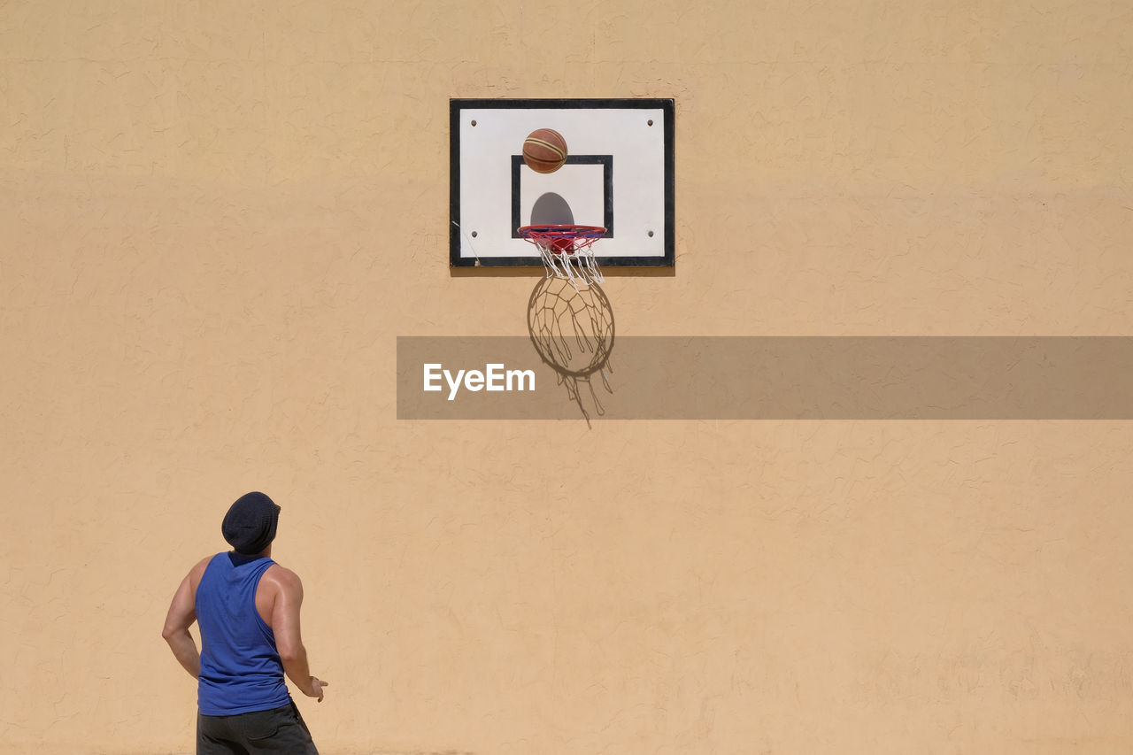 REAR VIEW OF MAN PLAYING WITH BASKETBALL AGAINST WALL