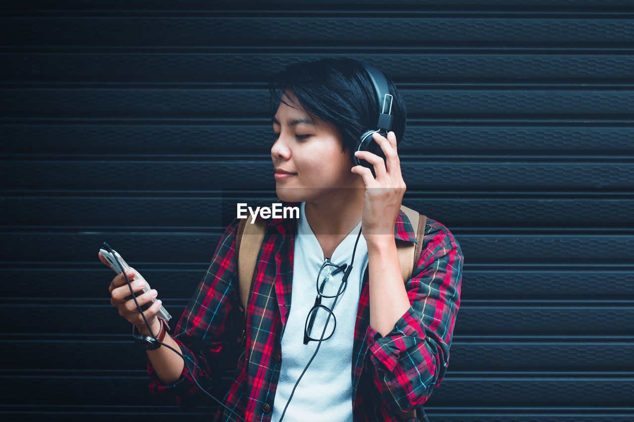 Smiling young woman listening music on headphones while standing against shutter