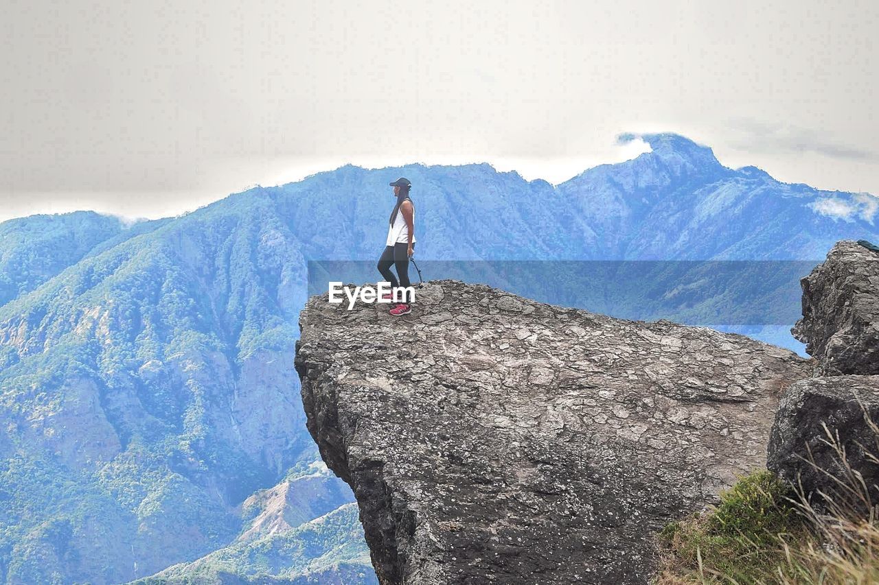 Low angle view of young woman standing on cliff by mountains