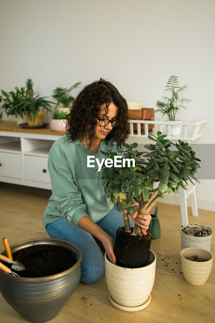 one person, potted plant, adult, plant, lifestyles, indoors, domestic life, women, houseplant, curly hair, growth, nature, hairstyle, flowerpot, brown hair, gardening, casual clothing, holding, female, domestic room, planting, young adult, home interior, looking, food and drink, standing, food, home, looking down, person, full length, day