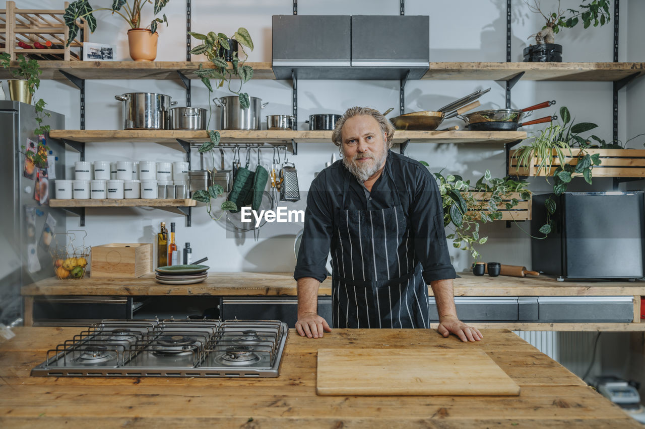 Confident mature chef staring while standing by kitchen counter in kitchen