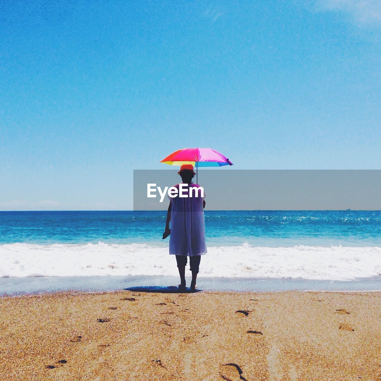 Full length rear view of woman with umbrella standing on beach against clear blue sky