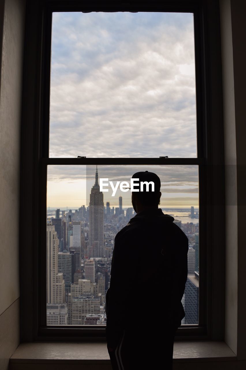 Rear view of man looking at empire state building amidst city through window