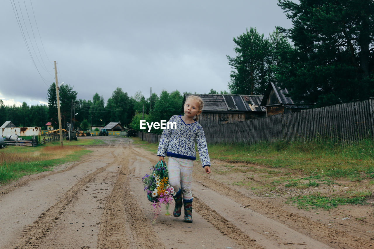 Girl in casual clothes walks through the village with a basket of wildflowers