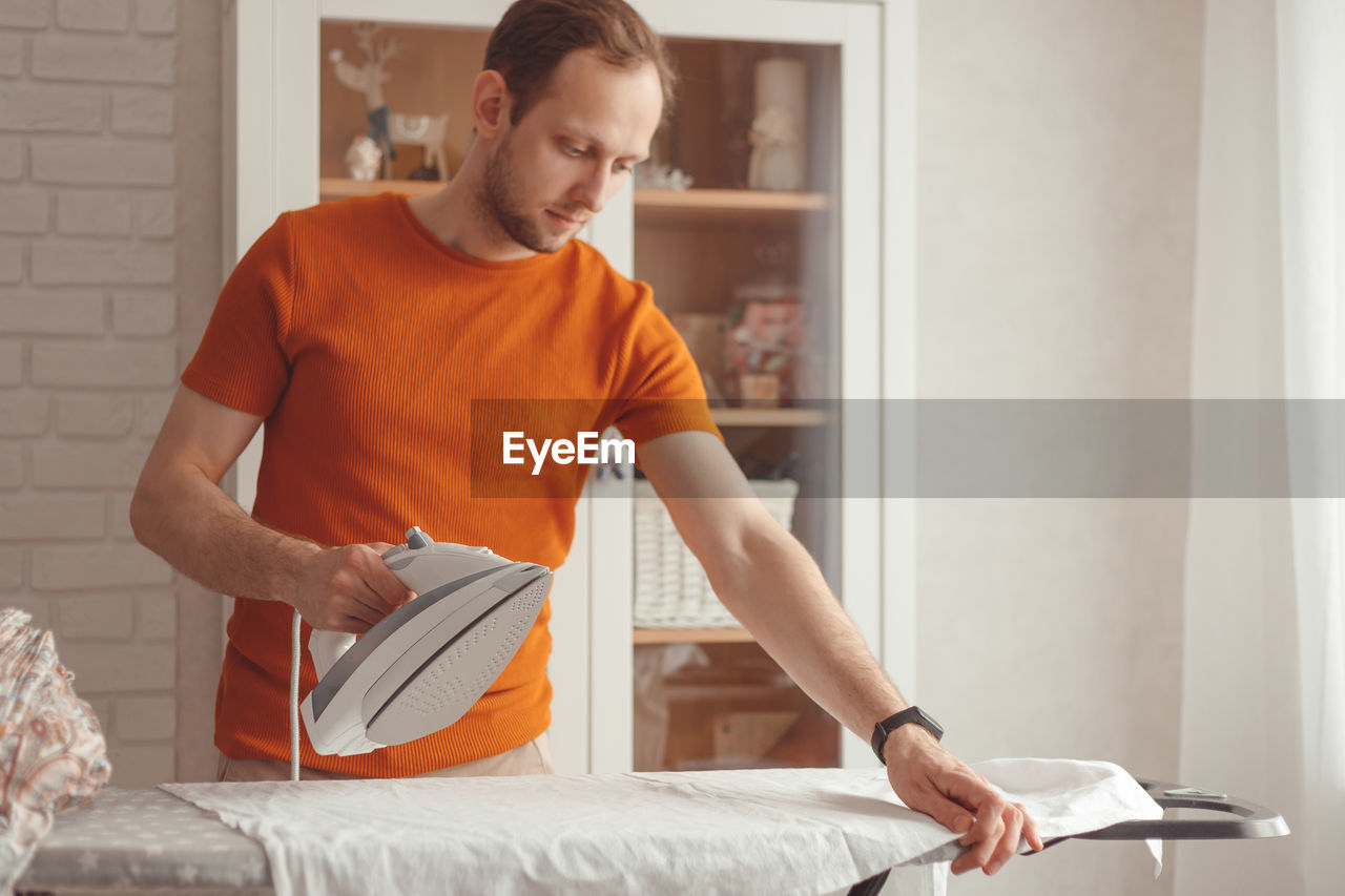 Young caucasian man ironing children's sheet on ironing board at home. men doing home chores