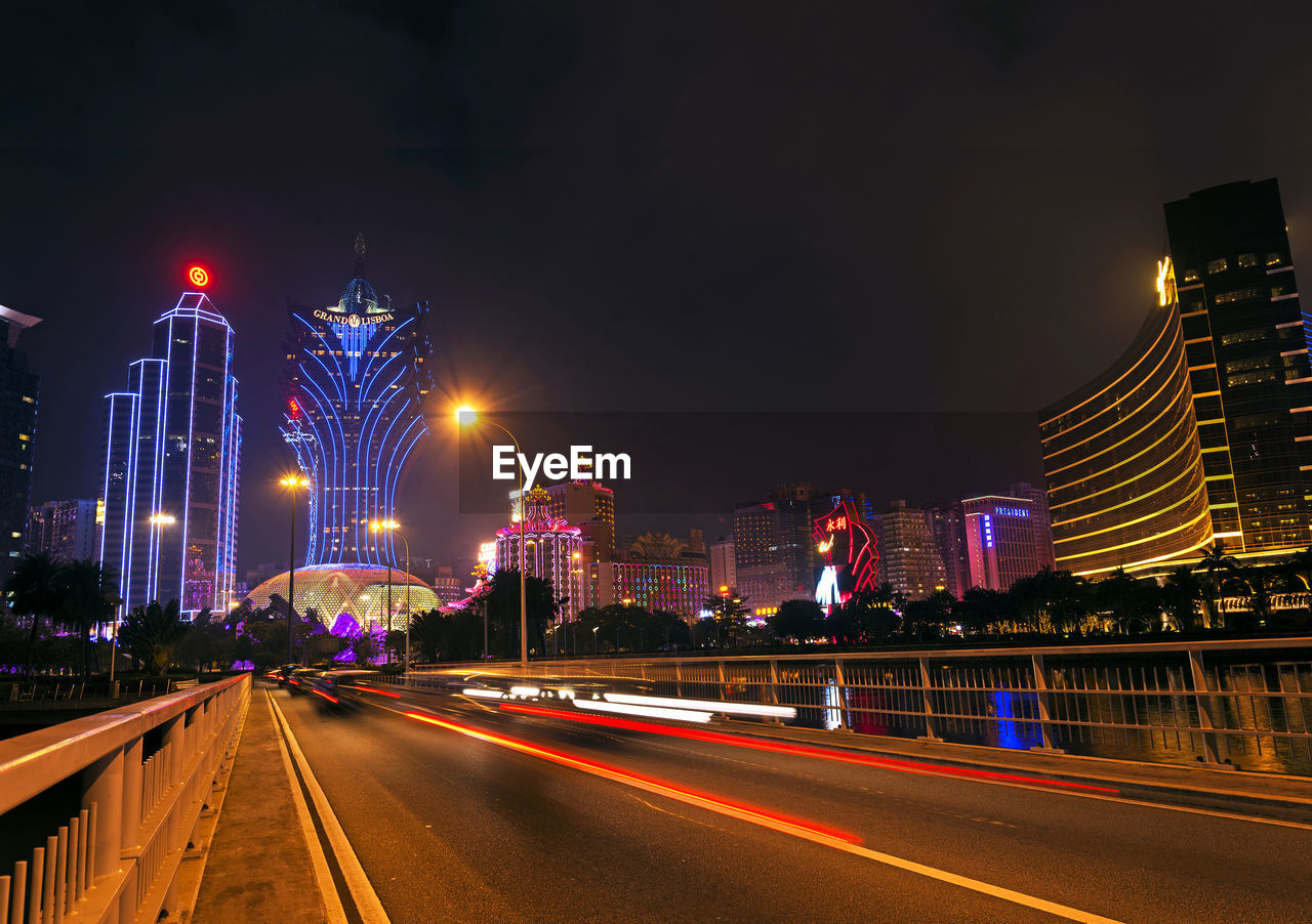 ILLUMINATED LIGHT TRAILS ON STREET AMIDST BUILDINGS AGAINST SKY AT NIGHT