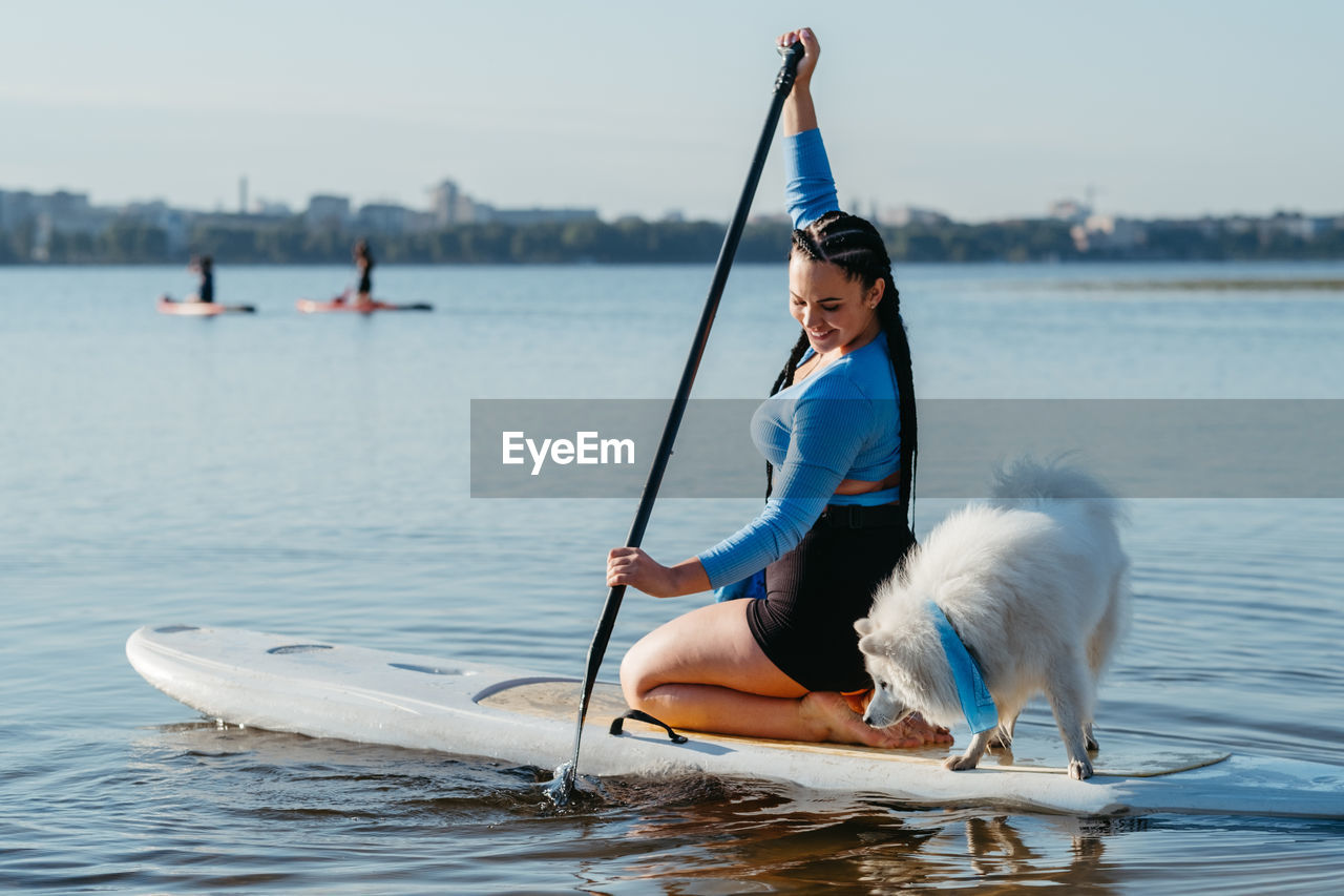 Woman with dreadlocks paddleboarding with dog snow-white japanese spitz on sup board on city lake